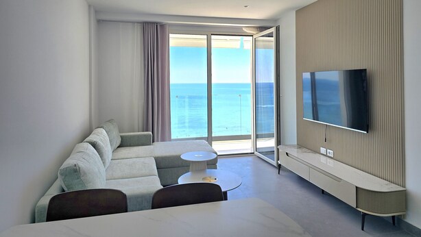 Quarto de hotel moderno com varanda ampla, sofá, mesa pequena e TV fixada na parede.