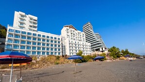 Um hotel à beira-mar com vários andares, uma praia arenosa em primeiro plano e diversos guarda-sóis.