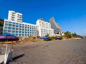 A beachfront hotel with multiple floors, a sandy beach in the foreground, and several umbrellas. - Panorama Deep Blue 001 (Shengjin)