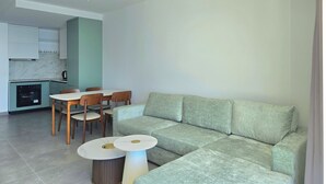 Uma sala de estar moderna com um sofá seccional, uma mesa de jantar com cadeiras e uma área de cozinha ao fundo.