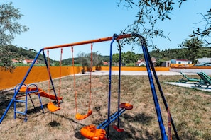 Un terrain de jeux avec des balançoires et un toboggan, entouré d’herbe et d’arbres.