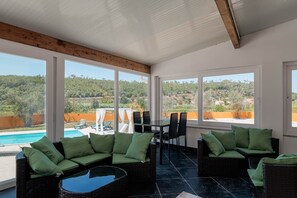 A sunlit room with large windows, wicker furniture, and a view of a pool and hills.