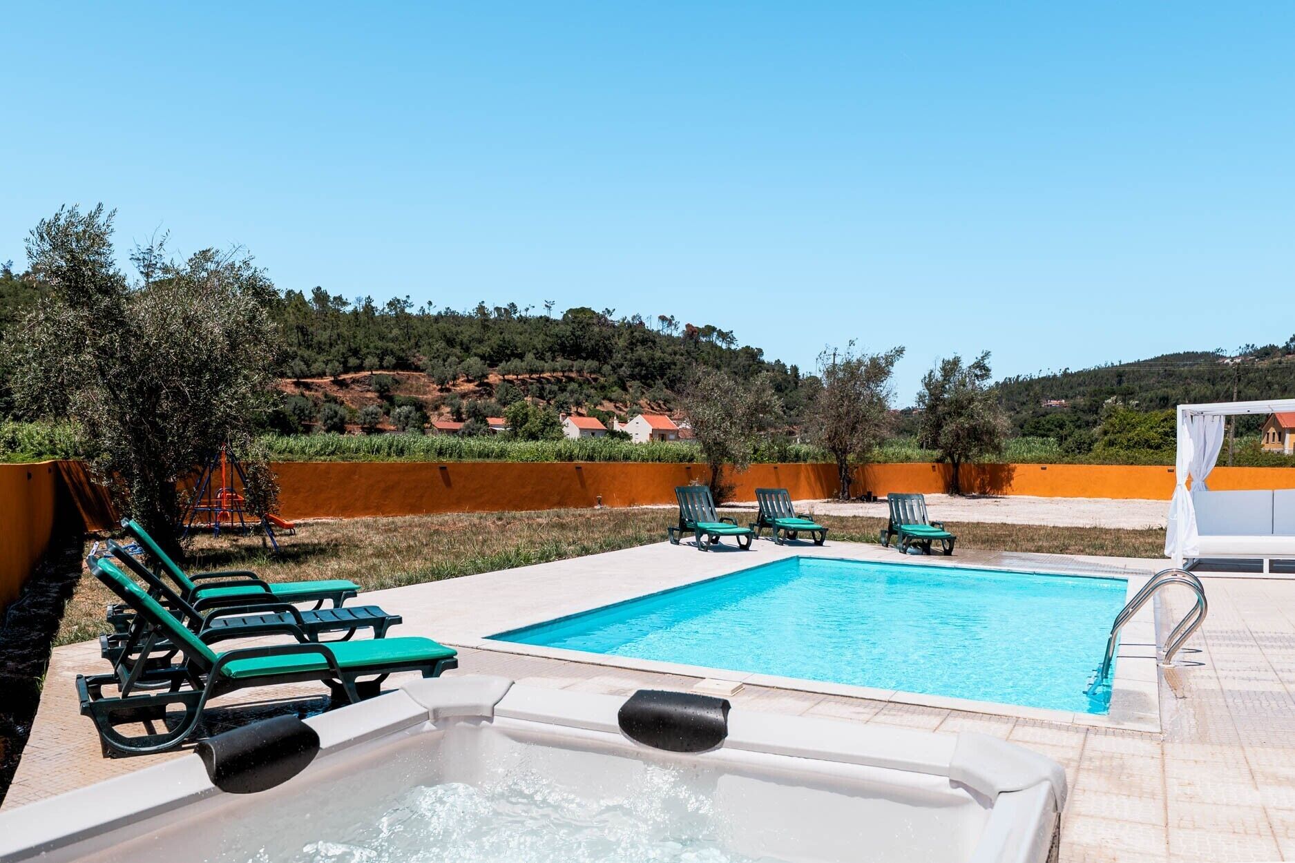 Una piscina con sillas de descanso y un jacuzzi, rodeada de jardines con árboles y vistas a una colina.