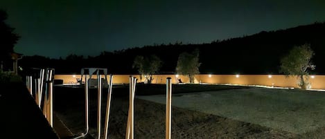 Una hilera de sillas metálicas con un área pavimentada al frente y un edificio iluminado al fondo.