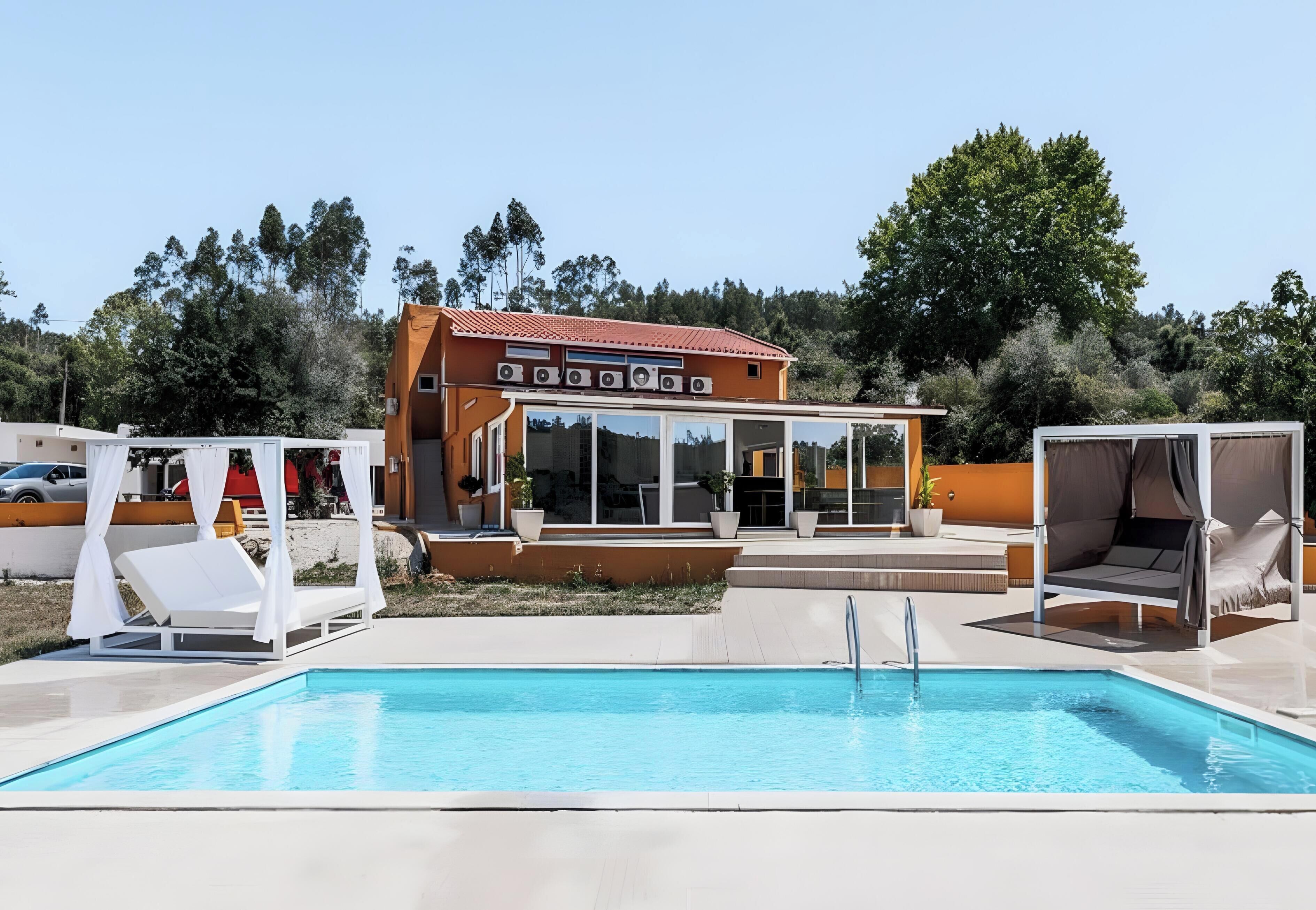 A swimming pool with lounge chairs and a covered seating area, surrounded by trees and a building with large windows.
