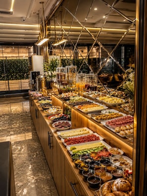 A buffet with various food items and beverages, a wooden counter, and a decorative ceiling structure. - MALABADİ HOTEL (Mersin)