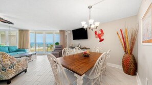 Ein geräumiger Wohnbereich mit einem Holzespeisetisch, weißen Stühlen, einer Couch und einem Balkon mit Blick auf den Strand.