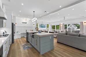 A modern kitchen with a central island, a dining area with a grey sofa, and a large window with a view of greenery.