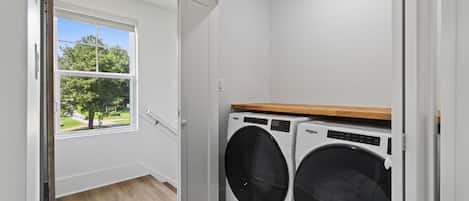 A laundry room with two front-loading washing machines, a window with a view of trees, and a wooden floor.