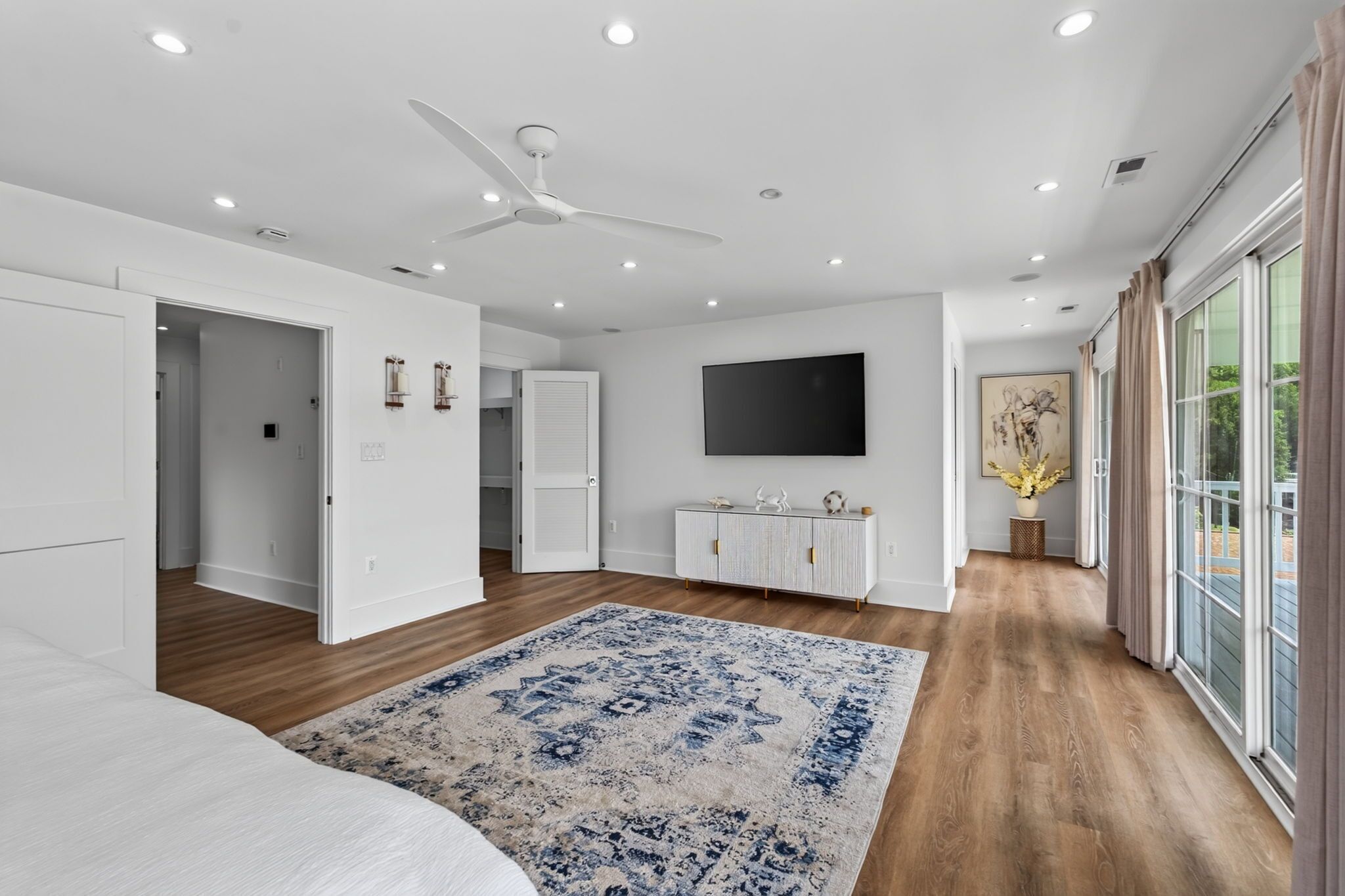 A modern bedroom with a large bed, a ceiling fan, a flat-screen TV, and a large window with curtains.