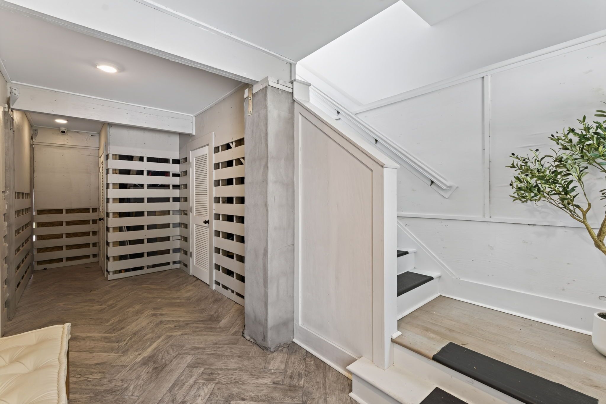 Un couloir avec un plancher en bois, une porte blanche dotée d’une fenêtre et un escalier agrémenté d’une plante en pot.