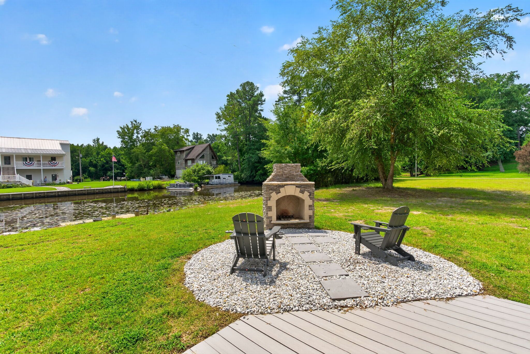 A deck with two Adirondack chairs and a stone fire pit, surrounded by a grassy area and a body of water.
