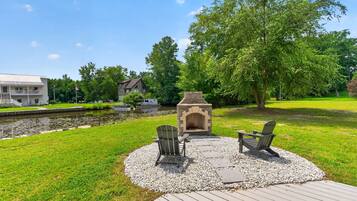 A deck with two Adirondack chairs and a stone fire pit, surrounded by a grassy area and a body of water.