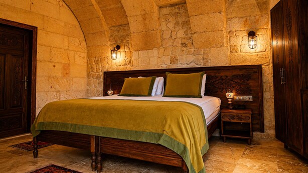 A room with a stone wall, a bed with a green bedspread, two wall-mounted lamps, and a wooden wardrobe. - La Mar Cave Cappadocia (Ürgüp)