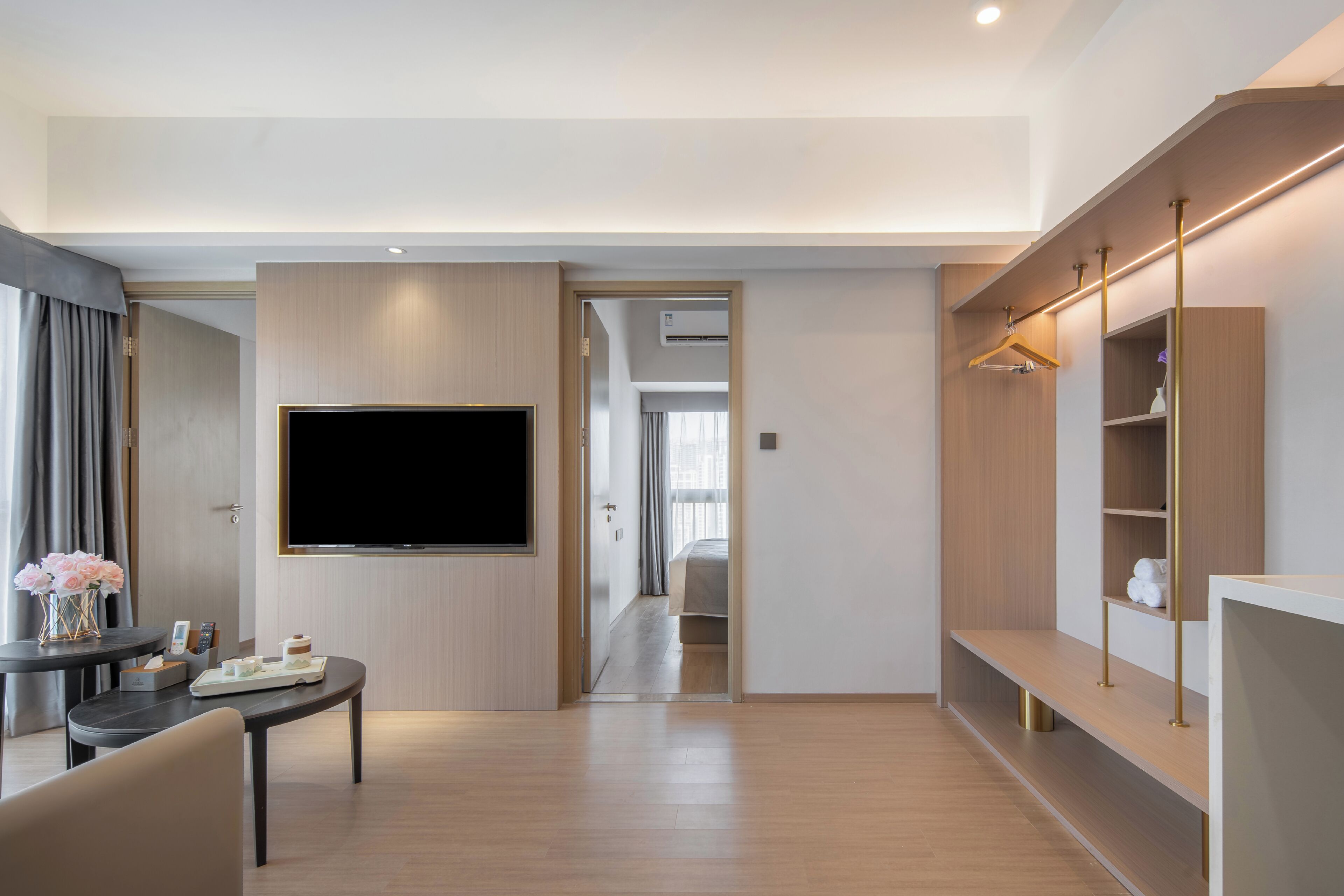 Una habitación de hotel moderna con un televisor de pantalla plana montado en la pared, un área de comedor con mesa y sillas, y un vestidor con estantes y una barra para colgar ropa.