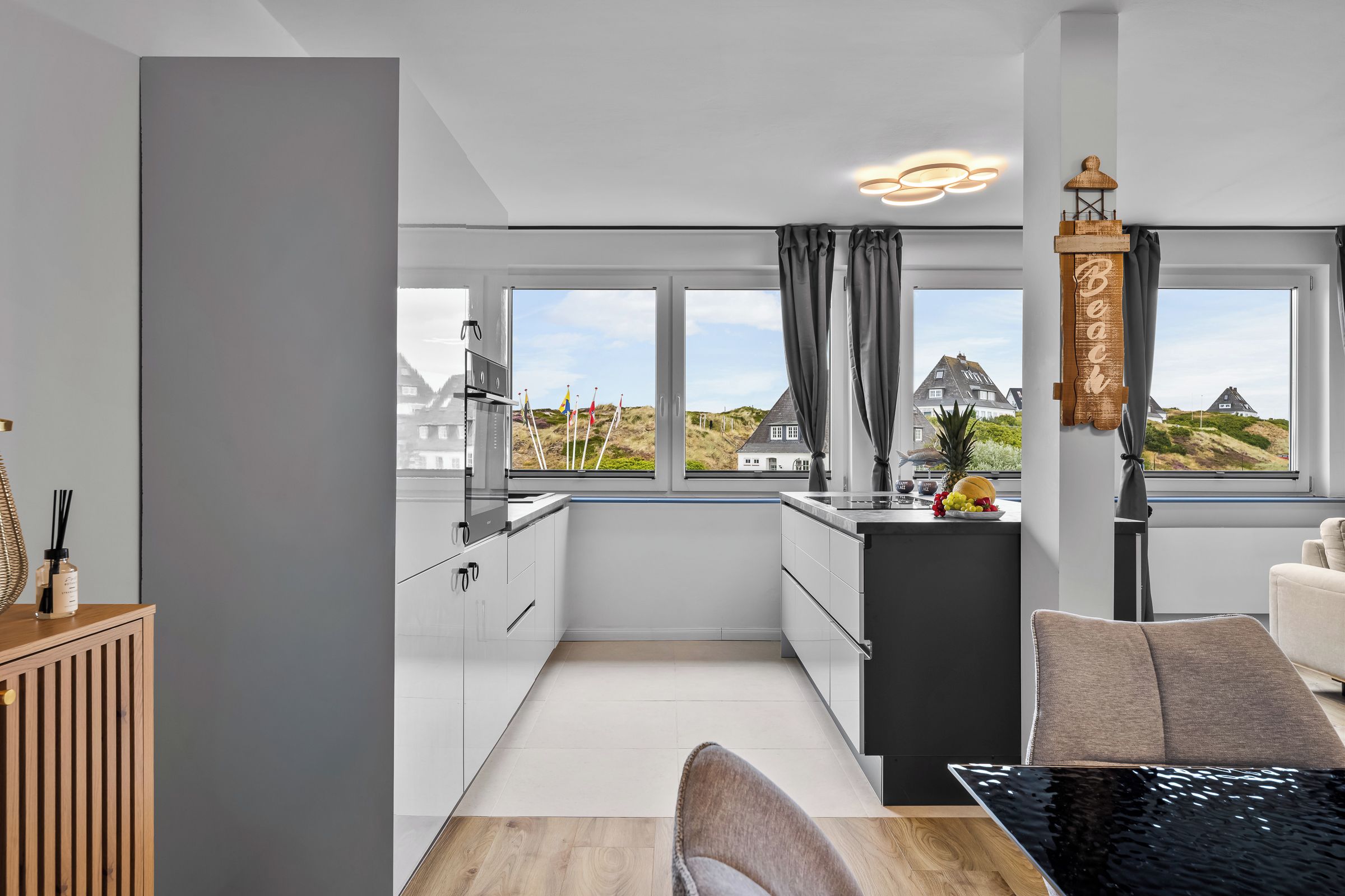 Et moderne køkken med udsigt til huse og klitter, et spiseområde med bord og stole, og et træskab med en flaske.