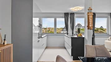 Et moderne køkken med udsigt til huse og klitter, et spiseområde med bord og stole, og et træskab med en flaske.