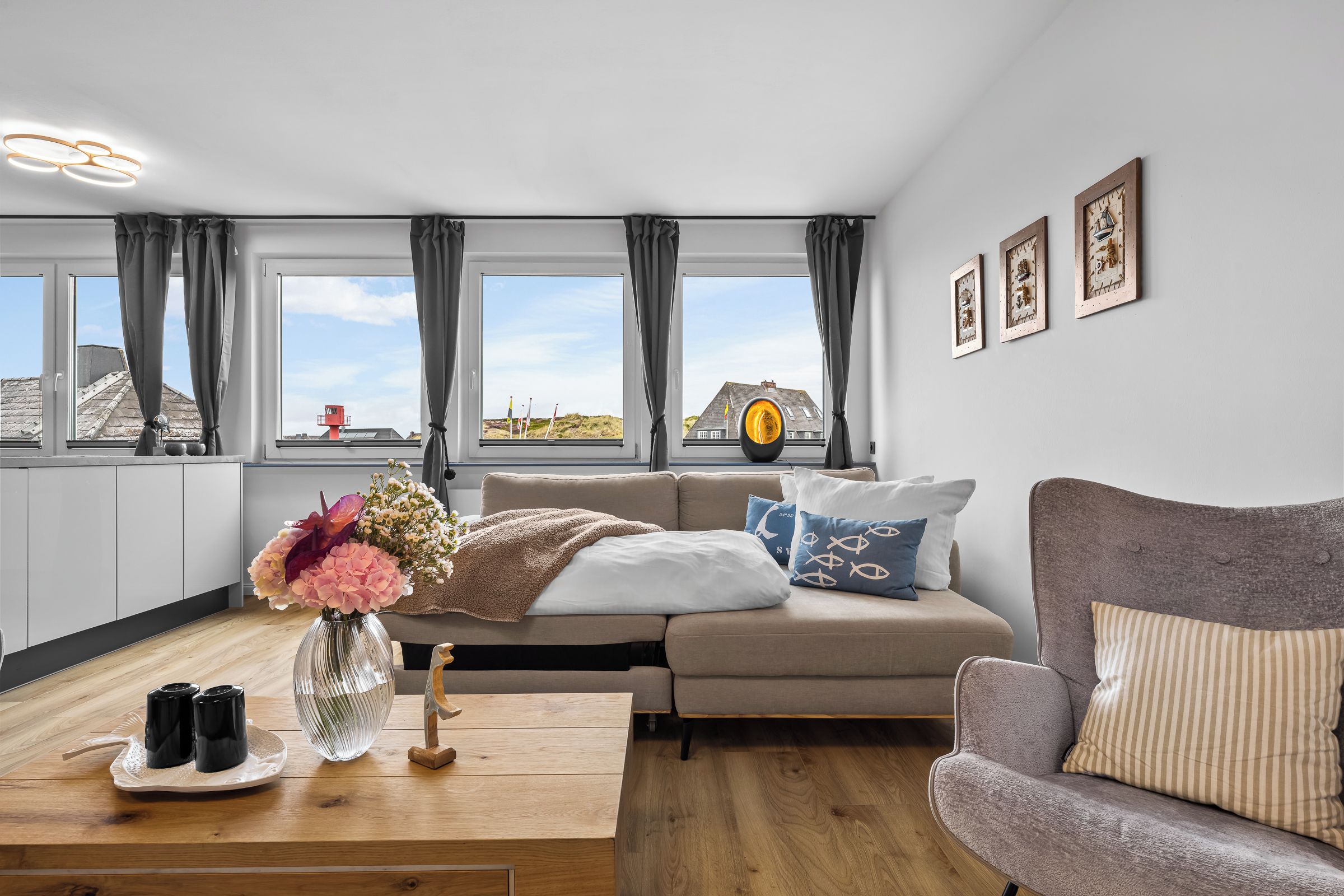 Ein modernes Wohnzimmer mit einer Couch, einem Holztisch mit Salzstreuern, einer Vase mit Blumen und Blick auf den Strand durch große Fenster.