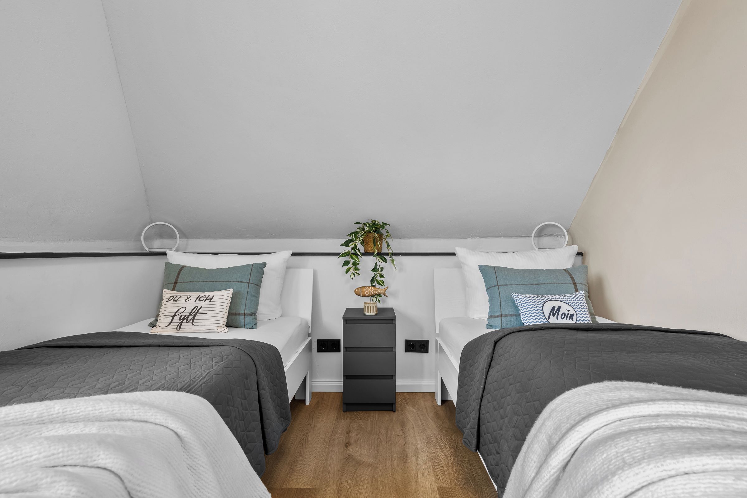 Ein Schlafzimmer mit zwei Einzelbetten, einem kleinen Nachttisch mit Pflanze und einem an der Wand befestigten Regal.