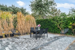 Un patio exterior con una mesa y sillas, rodeado de setos y pastos altos.