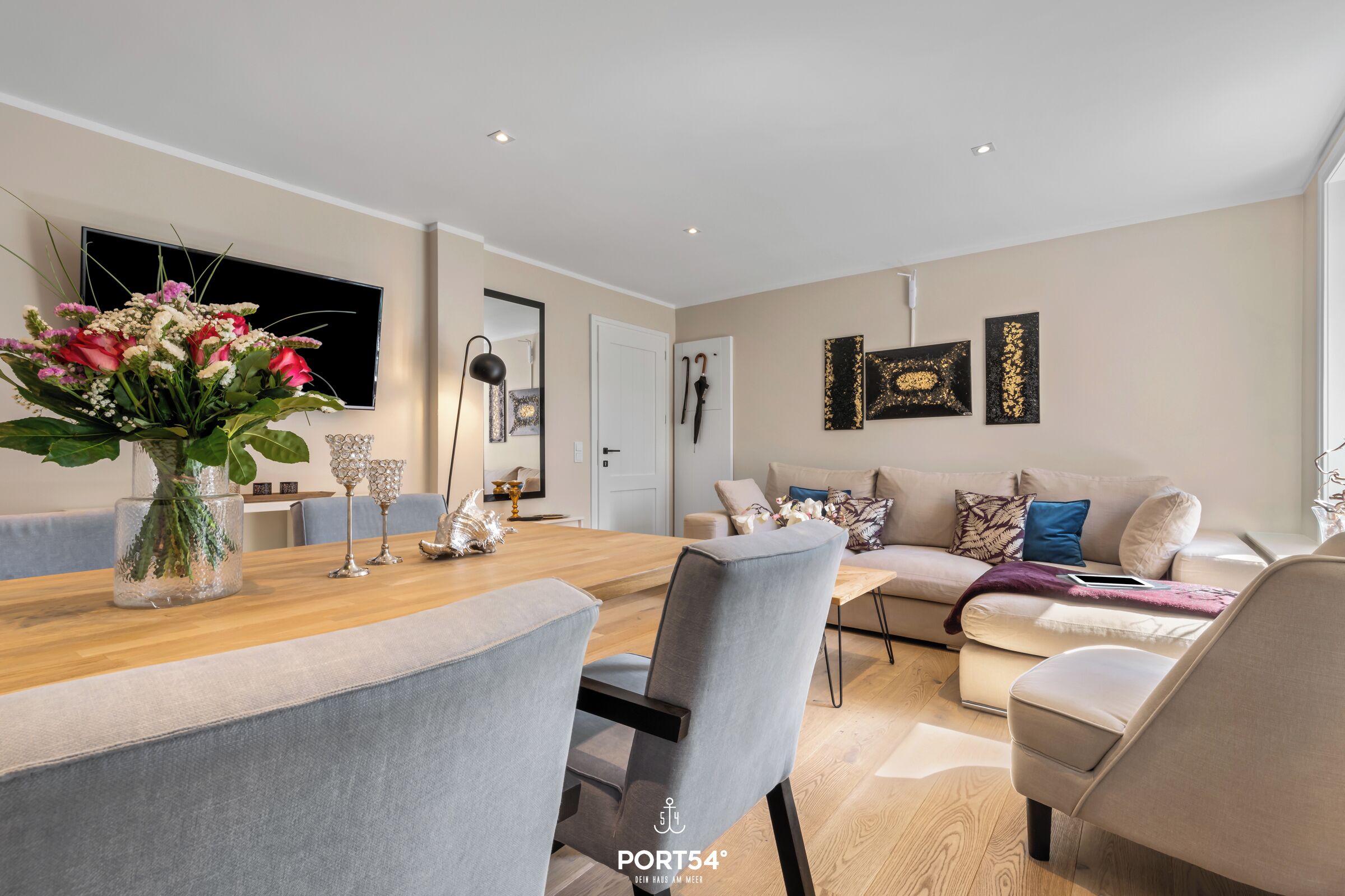 Een moderne woonkamer met een eethoek, met een houten tafel, grijs bekleden stoelen, een beige bank en een groot raam.