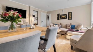 Un salon moderne avec un coin repas, comprenant une table en bois, des chaises à revêtement gris, un canapé beige et une grande fenêtre.