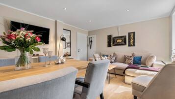Een moderne woonkamer met een eethoek, met een houten tafel, grijs bekleden stoelen, een beige bank en een groot raam.
