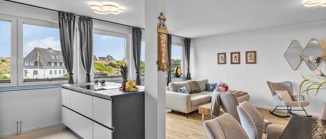 A modern kitchen with a large window, a dining area with a wooden table, and a living room with a sofa and chairs.