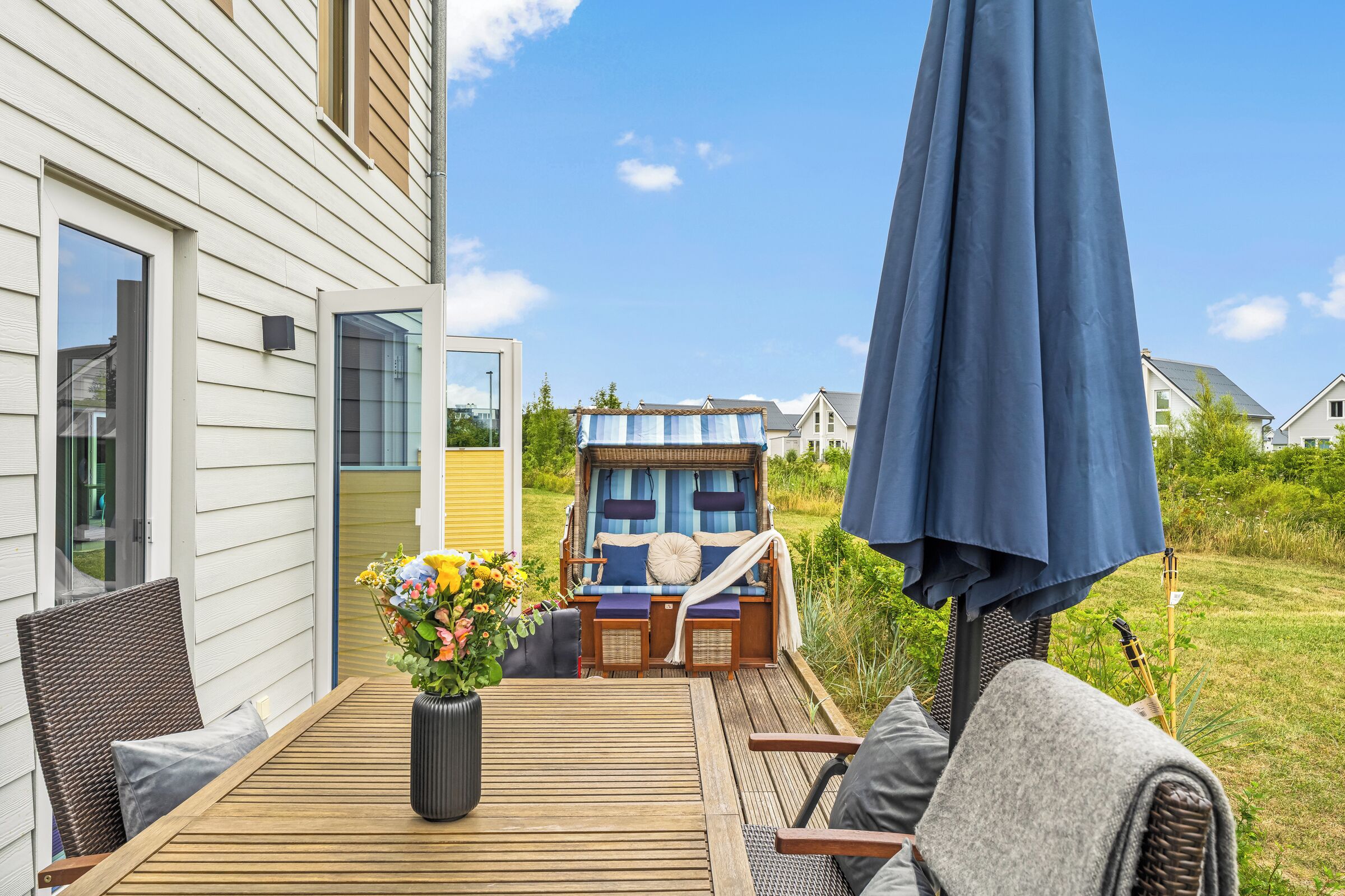 A deck with outdoor furniture, a wicker chair, a table with a vase of flowers, and a blue umbrella.