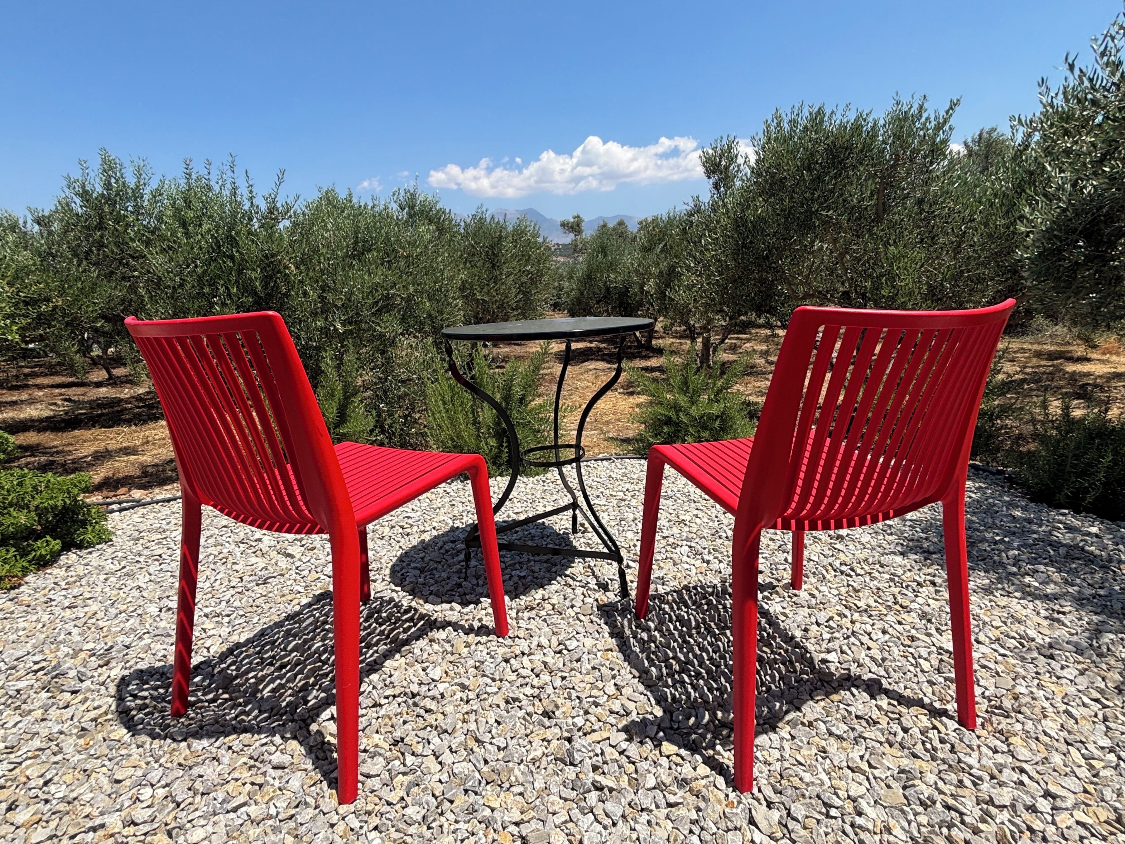 Twee rode stoelen en een kleine tafel op een grindpad, met olijfbomen op de achtergrond.