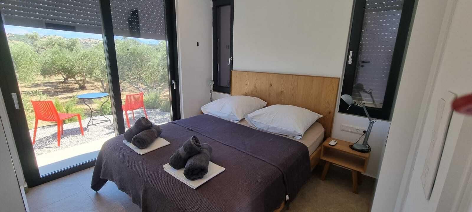 A bedroom with a bed, two pillows, a nightstand, a lamp, and a view of an outdoor area with a table and chairs.