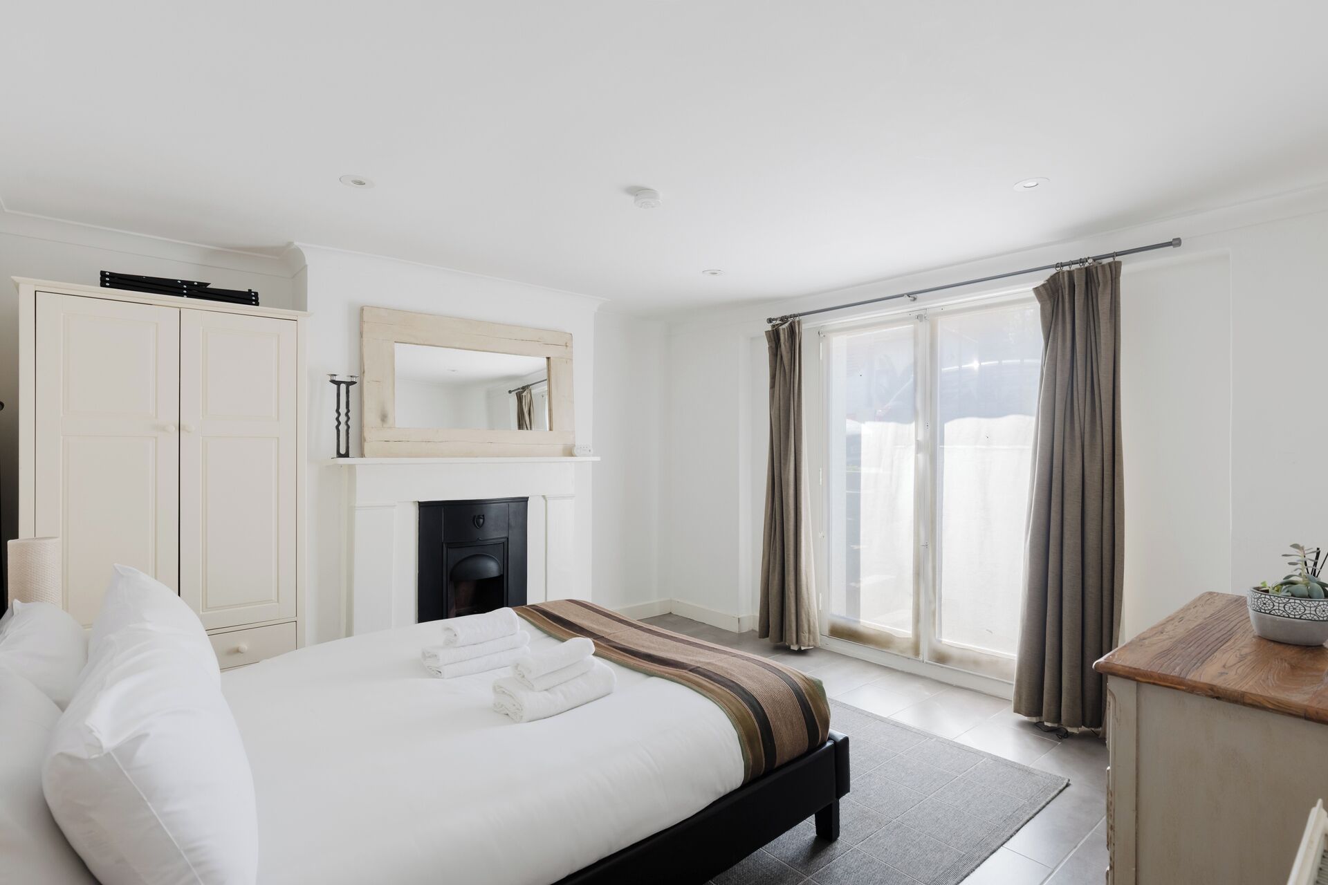 A hotel room with a bed, a fireplace, a wardrobe, a mirror, and a view of the outdoors.