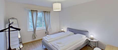 Une chambre à coucher comprenant un lit, une table de chevet, une lampe, un luminaire suspendu, une fenêtre avec des rideaux et une armoire.