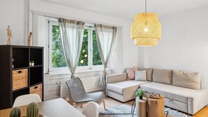 A modern living room with a sofa, a dining table, and a chair. There is a window with curtains, a hanging light fixture, and a bookshelf. - Charming 3 Bedroom Retreat (Zürich)