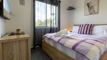 A bedroom with a bed, a bedside table, a window with curtains, a wall-mounted TV, and a framed picture.