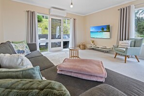 Ein modernes Wohnzimmer mit einem grauen Sofa, einem hellblauen Sessel, einer rosafarbenen Decke und einem Fernseher, der eine tropische Strandlandschaft zeigt.