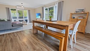 Una sala moderna con una mesa de comedor de madera, un banco y sillas blancas. Hay un sofá gris y una mesita auxiliar con una planta.