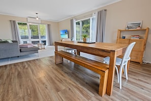 Ein modernes Wohnzimmer mit einem Holzesstisch, einer Bank und weißen Stühlen. Ein graues Sofa und ein kleiner Beistelltisch mit einer Pflanze sind ebenfalls vorhanden.
