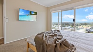 Un dormitorio con un ventanal grande, una cama con una colcha, un televisor instalado en la pared y piso alfombrado.