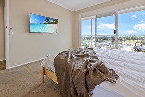 Ein Schlafzimmer mit großem Fenster, einem Bett mit Decke, einem an der Wand montierten Fernseher und Teppichboden.
