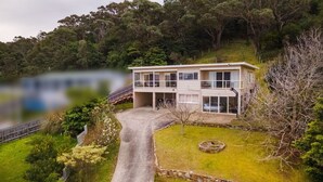 Una casa de dos pisos con un balcón, rodeada de abundante vegetación y un lago.