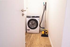 A compact laundry area with a front-loading washing machine, a vacuum cleaner, and a door. - Tide Resort 2a DG - Sauna. Balcony. Meernah. (Tossens)