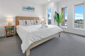 A bedroom with a large bed, a nightstand with a lamp, a painting on the wall, and a view through the window. - Coast and Canopy is the perfect place to slow down and reconnect (Eagle Point)