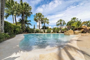 A swimming pool with clear water, surrounded by palm trees and rocks, with buildings in the background. - 2BR Pool-view apartment at Mirage Resort (Alexandra Headland)
