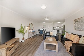 A modern living room with a flat-screen TV, wooden coffee table, and a dining area with chairs and a table. - 2BR Pool-view apartment at Mirage Resort (Alexandra Headland)