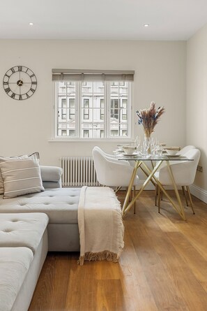 A modern living room with a glass dining table, a sofa, and a large window with a view of buildings. - Marble Arch's Gem - Large 2Bed 2Bath (London)