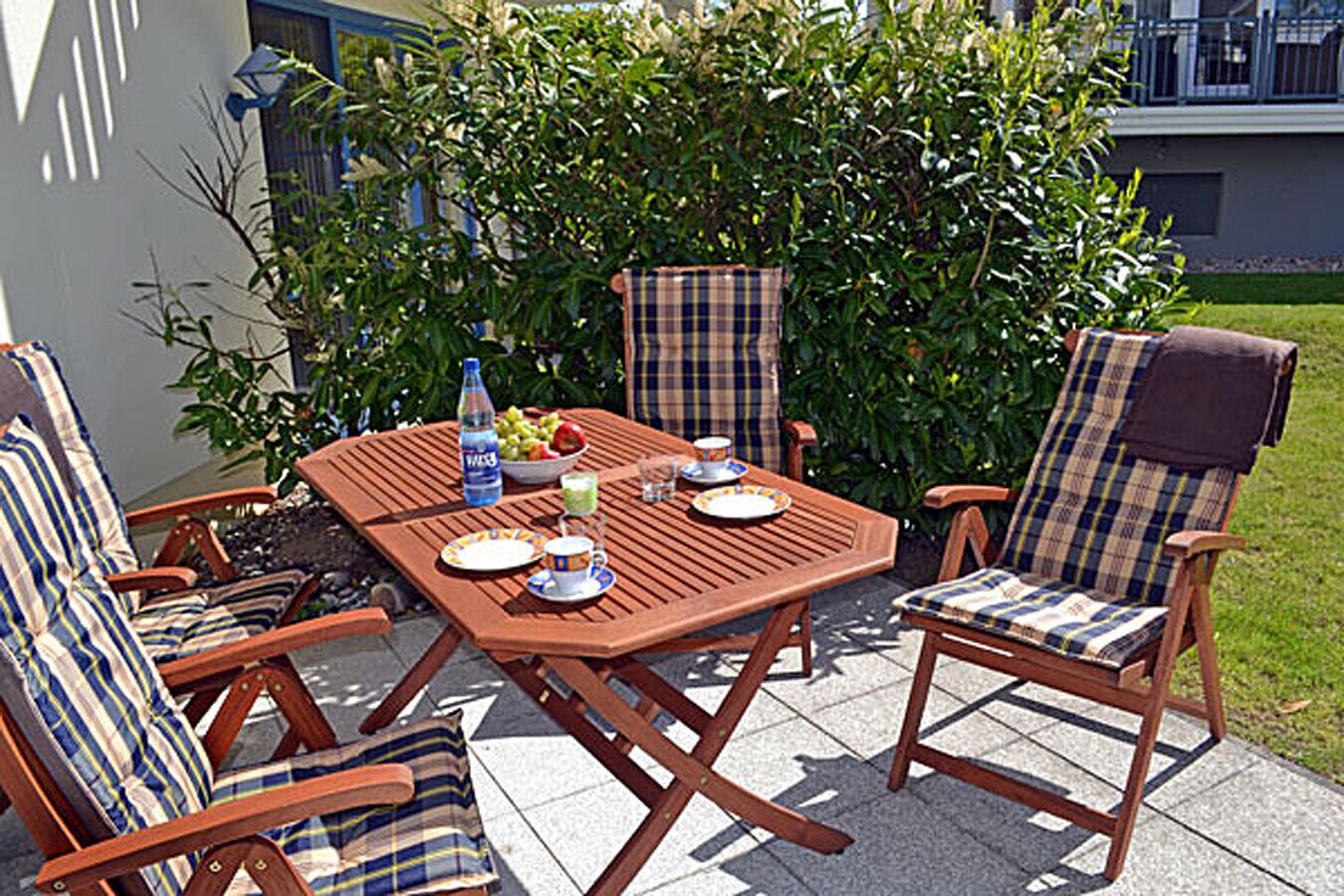 Eine Terrasse im Freien mit einem Holztisch, gedeckt für zwei Personen, umgeben von Stühlen mit Karomuster.