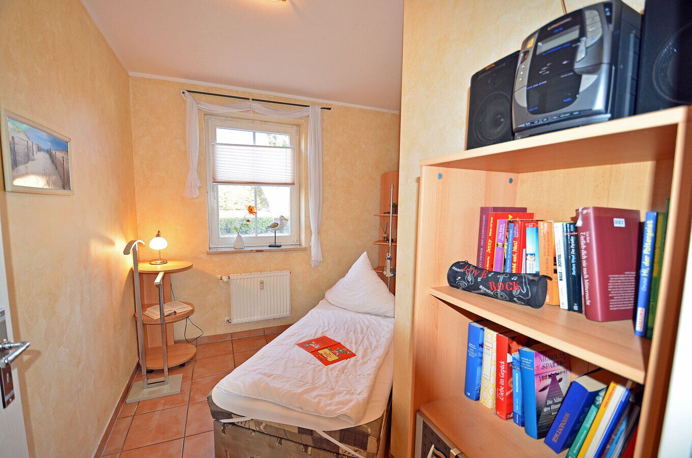 Ein kleines Schlafzimmer mit einem Einzelbett, einem Nachttisch, einer Lampe, einem Heizkörper, einem Fenster mit Vorhängen und einem Bücherregal mit Büchern.
