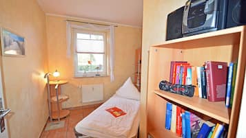 Ein kleines Schlafzimmer mit einem Einzelbett, einem Nachttisch, einer Lampe, einem Heizkörper, einem Fenster mit Vorhängen und einem Bücherregal mit Büchern.