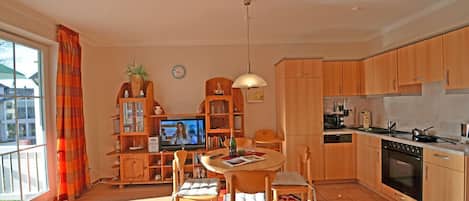 A well-lit, open-plan living and dining area with a kitchen, a television, and a dining table with chairs.